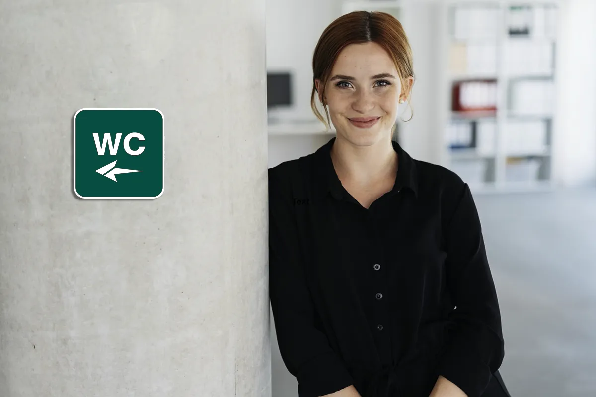 Ohne Angst zur/auf die Toilette Eine weibliche Person steht gegen eine Wand gelehnt neben einem grünen WC-Schild mit einem Pfeil nach links