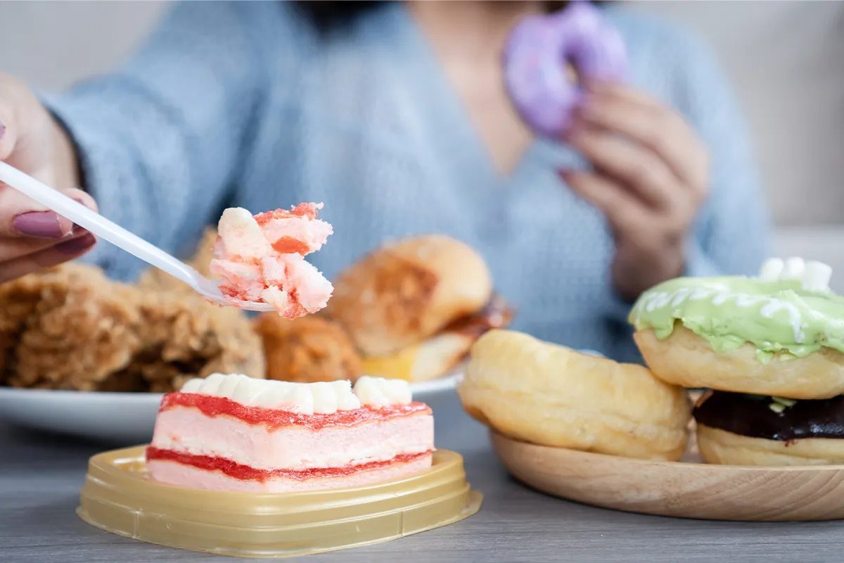 Eine weibliche verschwommene Person im Hintergrund mit angebissenem Donut in der einen Hand und Gabel mit einem anderen Kuchen in der anderen Hand. Davor stehen mehrere Teller mit Kuchen, Donuts und anderem Süssgebäck