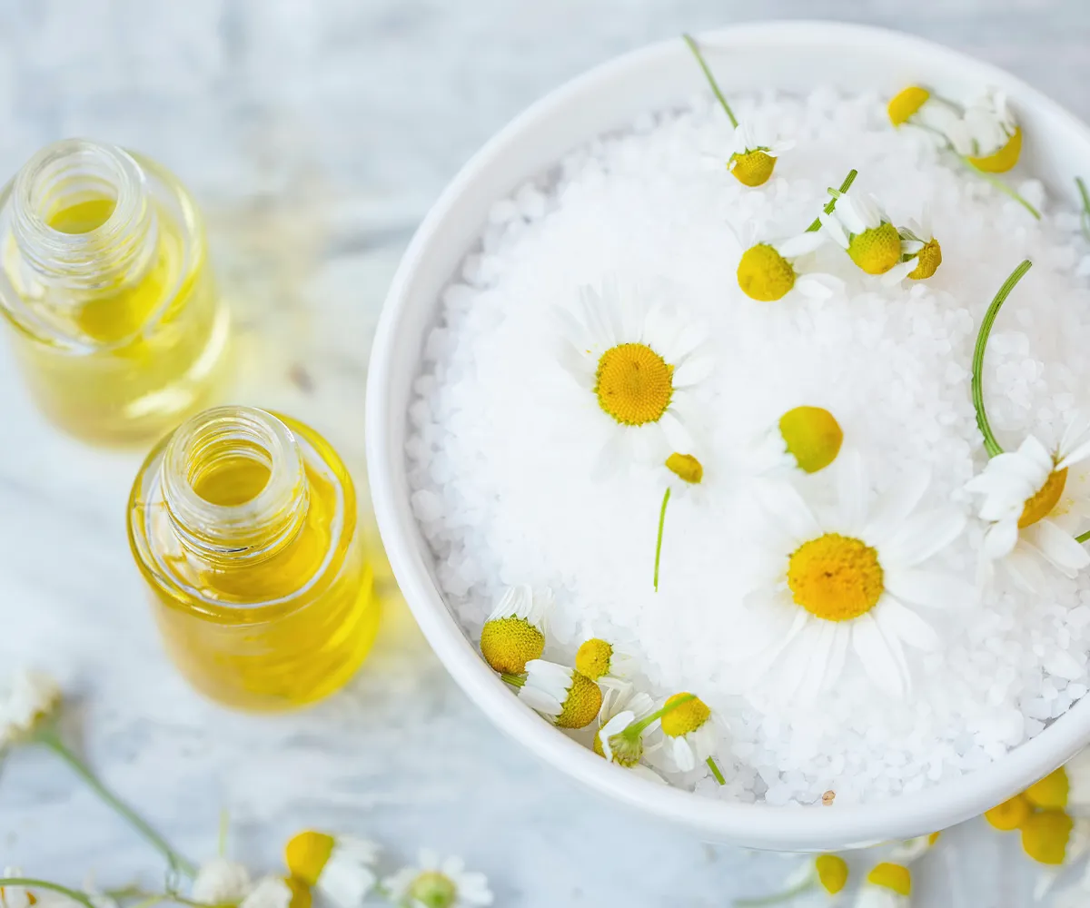 Kamilleblüten auf einem weißen Grandulat in einer Schüssel. Daneben zwei offene Fläschchen mit einer gelblichen Flüssigkeit drinnen