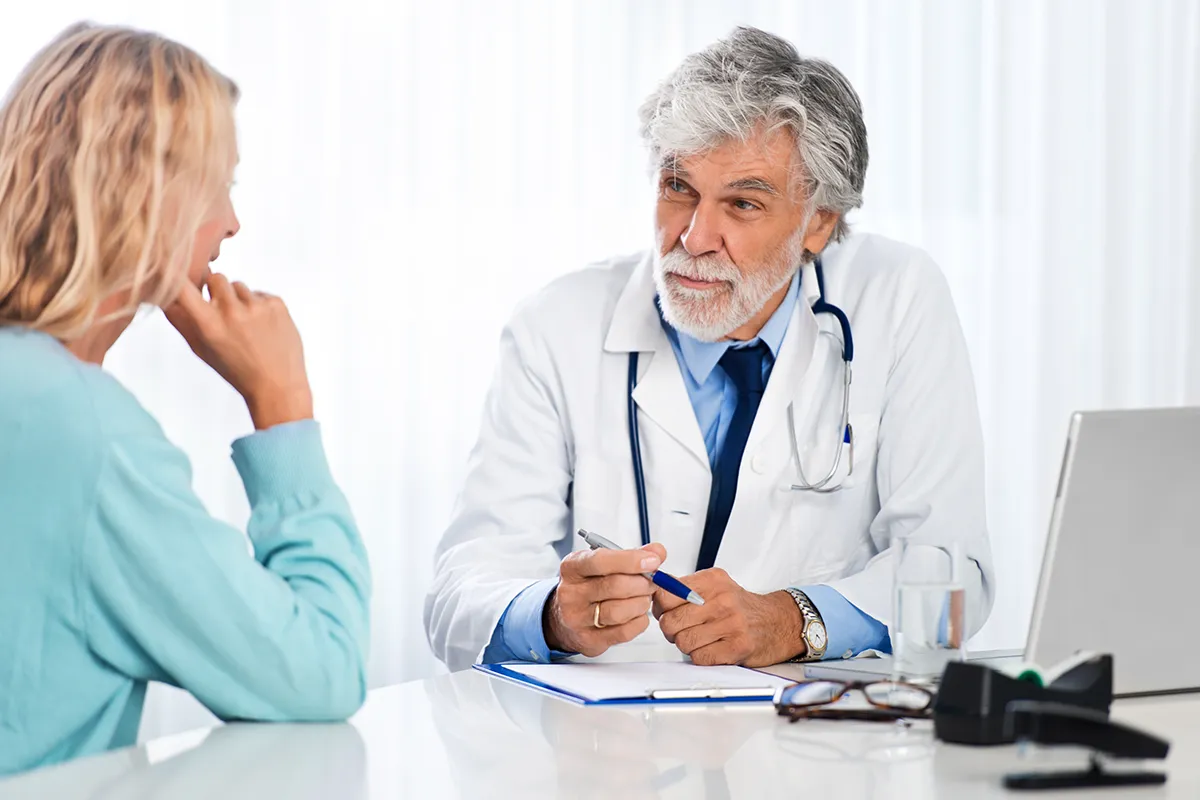 Eine weibliche Person sitzt an einem Tisch vor einem älteren medizinischen Personal mit Laptop und Klemmbrett vor sich