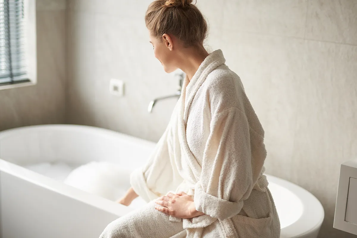 Eine Frau im Bademantel sitzt auf der Kante einer Badewanne, dadrinnen sind viele Seifenblasen zu sehen