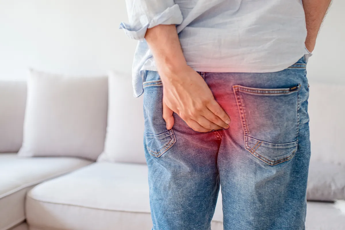 Eine männliche Person hält sich mit der linken Hand den Afterbereich. Schemenhaft ist der Bereich rot überlagert