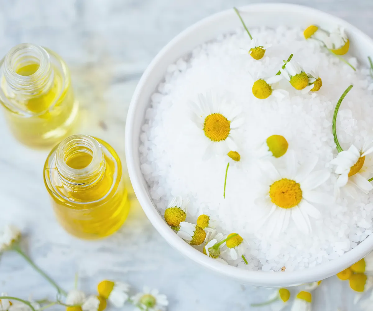 Schmerzen beim Stuhlgang Behandlungsmöglichkeiten Kamilleblüten auf einem weißen Grandulat in einer Schüssel. Daneben zwei offene Fläschchen mit einer gelblichen Flüssigkeit drinnen
