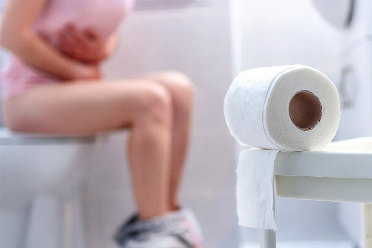 Schmerzen beim Stuhlgang Lidoproct lindert Eine weibliche Person gekrümmt auf einer Toilette sitzend im Hintergrund mit einer Rolle Klopapier im Vordergrund