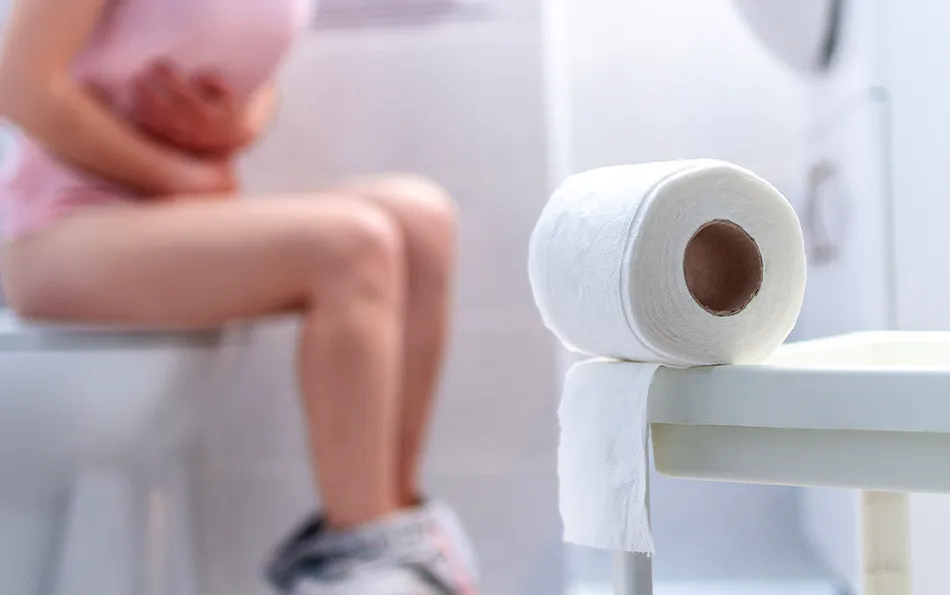 Eine weibliche Person gekrümmt auf einer Toilette sitzend im Hintergrund mit einer Rolle Klopapier im Vordergrund