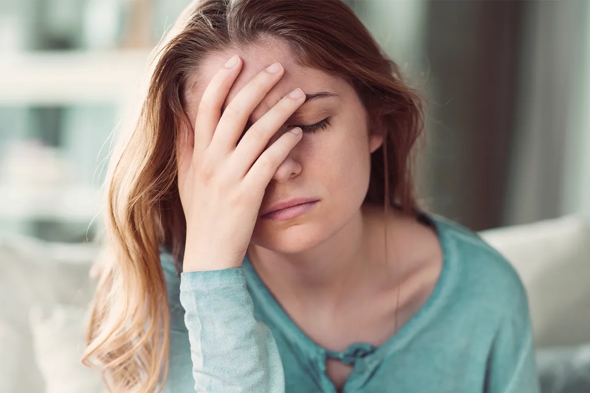Eine niedergeschlagen dreinschauende weibliche Person mit geschlossenen Augen und einer Hand vor dem halben Gesicht