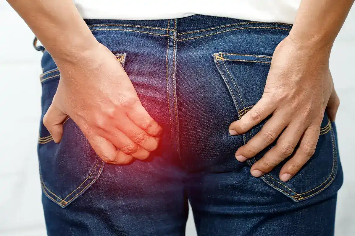 Eine männliche Person hält sich sehr verkrampft mit der linken Hand die linke Pobacke. Schemenhaft ist der Bereich rot überlagert